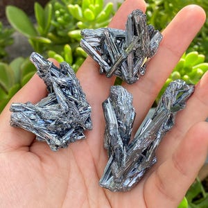 Pode incluir: Três grupos de cristais de estibnite metálicos cinza-prateados com uma estrutura prismática semelhante a agulhas. Cada peça exibe uma superfície lustrosa e altamente reflexiva com formações cristalinas afiadas e alongadas.