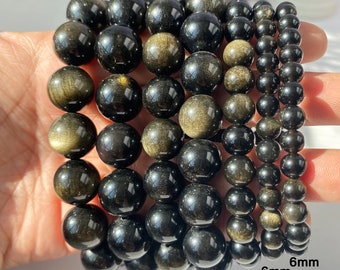 Könnte beinhalten: Sieben Armbänder mit goldenen Obsidianperlen in verschiedenen Größen, von 6 mm bis 14 mm. Die Perlen sind schwarz und goldfarben, mit einer polierten, reflektierenden Oberfläche. Die Armbänder werden nebeneinander gezeigt.