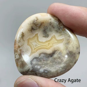 May include: A close-up of a smooth, polished, light brown agate stone with a swirling pattern of white, gray, and yellow. The stone is called "Crazy Agate".
