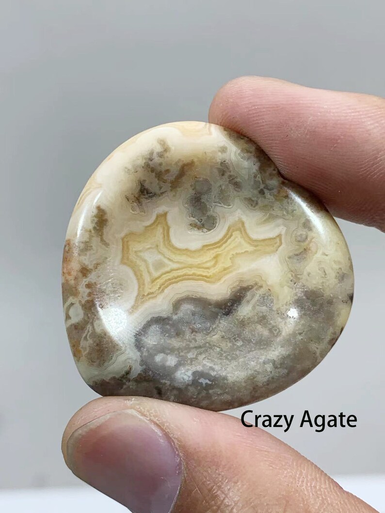 May include: A close-up of a smooth, polished, light brown agate stone with a swirling pattern of white, gray, and yellow. The stone is called "Crazy Agate".