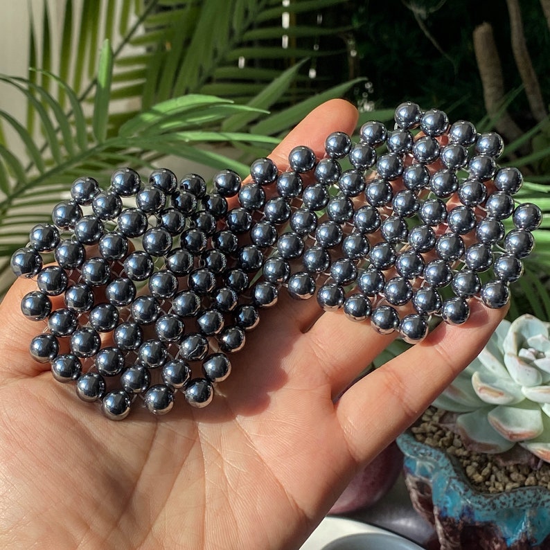 Pu&ograve; includere: Primo piano di una mano che tiene un grande gruppo di piccole perline metalliche rotonde di colore grigio scuro. Le perline sono disposte in uno schema geometrico, creando un design unico e accattivante.