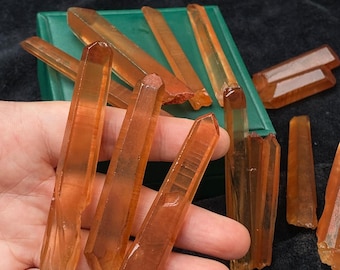 May include: Several orange crystal points, some held in a hand, others arranged near a green box. The crystals have a translucent quality and varying shapes. The background is a dark, textured fabric, providing contrast to the crystals' colour.
