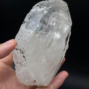 Op de afbeelding: Een groot, helder kwartskristal met een ruw, getextureerd oppervlak. Het kristal heeft iriserende gebieden, die een regenboog van kleuren weergeven. Het kristal wordt in een hand gehouden tegen een zwarte achtergrond.