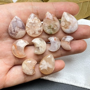 May include: A collection of polished gemstones in various shapes, including teardrops and crescent moons. The stones display a mix of colors, such as white, peach, and hints of brown, with unique patterns. The gemstones are held in a hand.