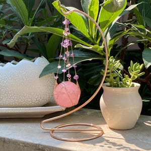 May include: A rose gold metal stand supports a pink crystal sphere, decorated with hanging amethyst and pink gemstone strands. The sphere is enclosed in a delicate chain design. A small white ceramic pot with a succulent plant is to the right.