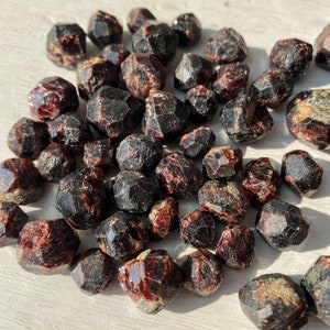 May include: A collection of dark red garnet crystals, some with a slight metallic sheen. The crystals are various sizes and shapes, with some being more rounded and others more angular.