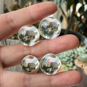 May include: Four clear glass spheres, each reflecting a different view of a room with plants. The spheres are arranged in a row on a hand.