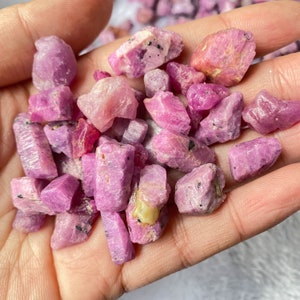 May include: A collection of pink ruby rough stones, some with a darker pink hue, in a hand. The stones are various sizes and shapes, with some having a more rectangular shape.