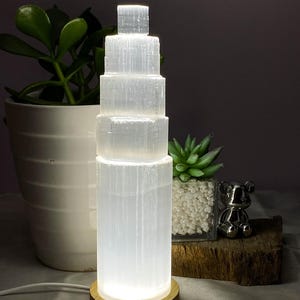 May include: A white selenite tower lamp with a wooden base. The lamp is composed of stacked rectangular blocks, illuminated from within. The text "M-(200x50mm,800g)" is visible at the bottom. The lamp is next to a potted plant.