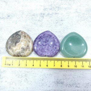May include: Three polished, heart-shaped stones in a row. The stone on the left is brown and white with a swirling pattern. The middle stone is purple and the stone on the right is green.