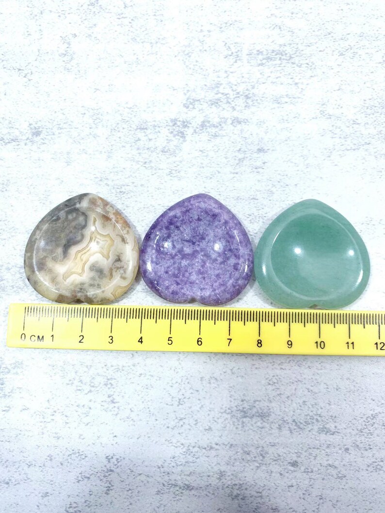May include: Three polished, heart-shaped stones in a row. The stone on the left is brown and white with a swirling pattern. The middle stone is purple and the stone on the right is green.