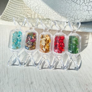Puede incluir: Cinco recipientes transparentes de plástico con forma de caramelo llenos de piedras preciosas de colores. Los recipientes están dispuestos en fila sobre una superficie de madera blanca. Las piedras preciosas son turquesa, roja, marrón, verde y amarilla.