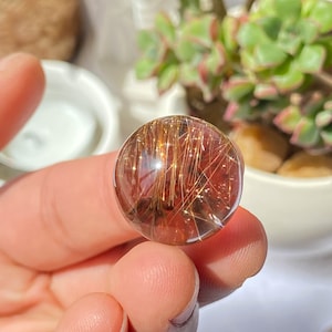 K&ouml;nnte beinhalten: Eine klare Quarzkugel mit goldenen Rutil-Einschl&uuml;ssen. Die Kugel hat einen Durchmesser von etwa 2,5 cm und eine durchscheinende, leicht r&ouml;tliche Farbe.