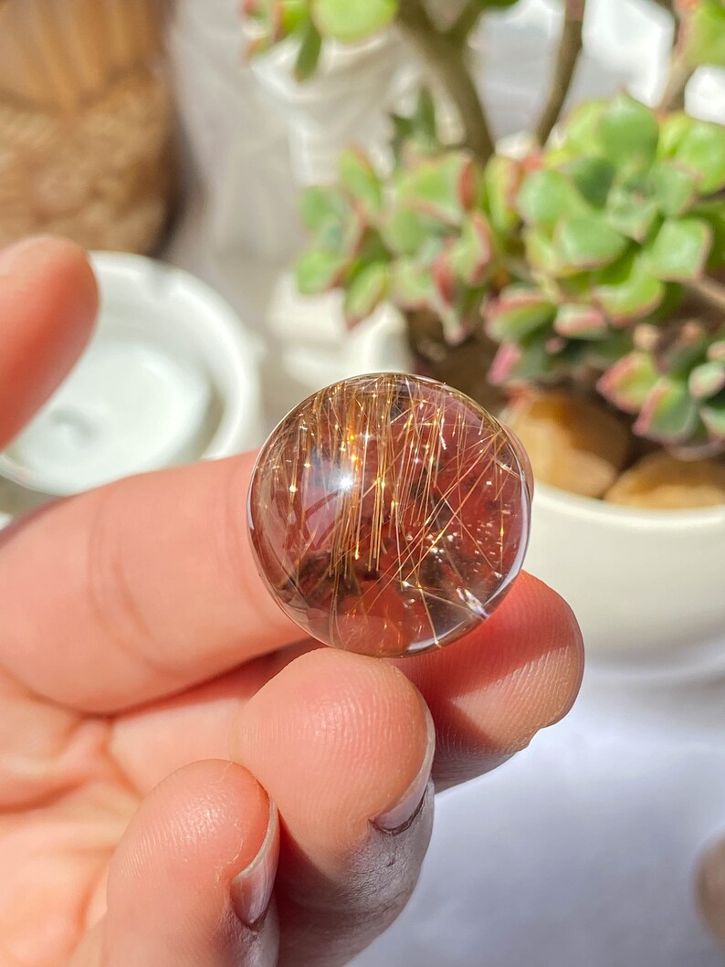 K&ouml;nnte beinhalten: Eine klare Quarzkugel mit goldenen Rutil-Einschl&uuml;ssen. Die Kugel hat einen Durchmesser von etwa 2,5 cm und eine durchscheinende, leicht r&ouml;tliche Farbe.