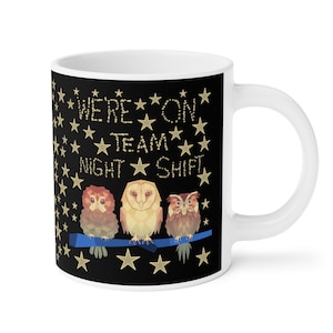 Puede incluir: Taza de cerámica blanca con un diseño negro que presenta tres búhos posados en una rama. El texto "We're on Team Night Shift" está escrito en estrellas doradas sobre un fondo negro.