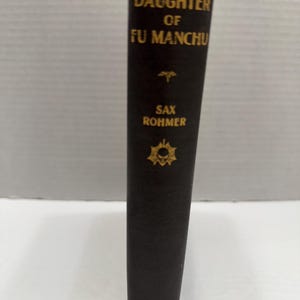 May include: A vintage hardcover book titled "Daughter of Fu Manchu" by Sax Rohmer. The book has a dark cover with gold lettering and decorative elements. The publisher's name, Collier, is printed at the bottom.