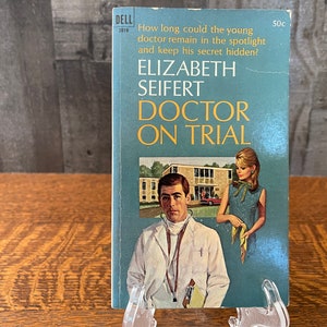 May include: A blue paperback book with the title "Doctor on Trial" by Elizabeth Seifert. The cover features a man in a white doctor's coat and a woman in a blue dress.