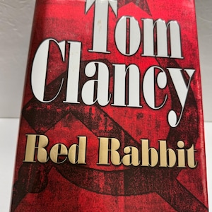 May include: A hardcover book titled "Tom Clancy Red Rabbit." The cover is predominantly red with white and gold lettering. The design includes a star and a hammer and sickle symbol.