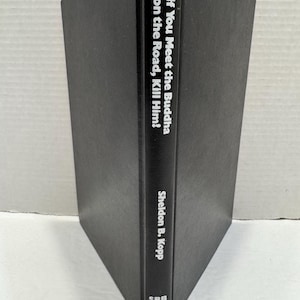 May include: A hardcover book with a black cover. The title "If You Meet the Buddha on the Road, Kill Him!" is printed vertically in white. The author's name, Sheldon B. Kopp, is also in white. The book has a small floral design at the bottom.