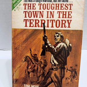 May include: Vintage paperback book cover for "The Toughest Town in the Territory" by Tom West. The cover features a cowboy with a gun, and other cowboys on horseback. The title is in large red letters.