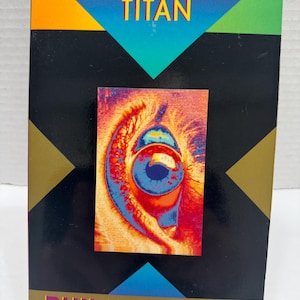 May include: A paperback book titled "THE GAME-PLAYERS OF TITAN" by Philip K. Dick. The cover features a colorful eye graphic and geometric shapes in orange, blue, and green. The book has a black background.