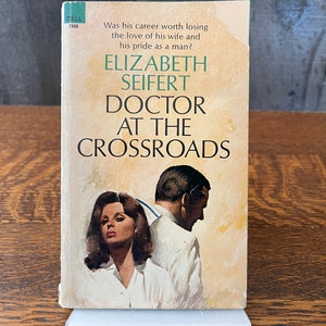 May include: A vintage paperback book cover with a yellow background and a couple in the background. The title is "Doctor at the Crossroads" by Elizabeth Seifert. The book was published in 1968.