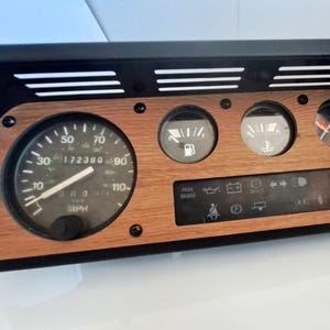 May include: A black dashboard with wood effect detailing. It includes a speedometer, fuel gauge, and other warning lights. The dashboard is rectangular with vents at the top. The speedometer reads in mph.