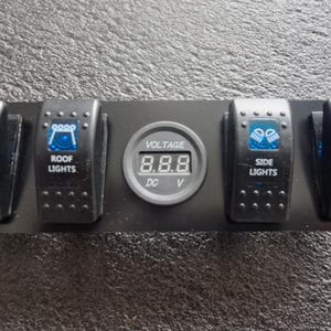 Op de afbeelding: Een zwart rechthoekig schakelpaneel met afgeronde uiteinden, voorzien van vier zwarte tuimelschakelaars en een centraal digitaal voltage-display. De schakelaars zijn gelabeld met LED LIGHT BAR, ROOF LIGHTS, SIDE LIGHTS en SPOT LIGHTS met blauwe iconen.