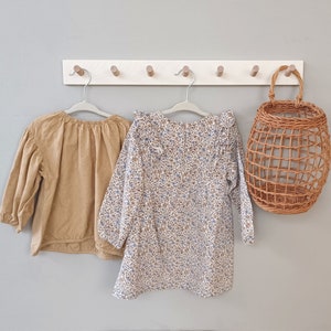 Op de afbeelding: Twee kindertops en een geweven mand hangen aan een muur. De ene top is bruin met een oogjespatroon, de andere is een jurk met bloemenprint met blauwe en bruine bloemen. De mand is gemaakt van geweven riet.