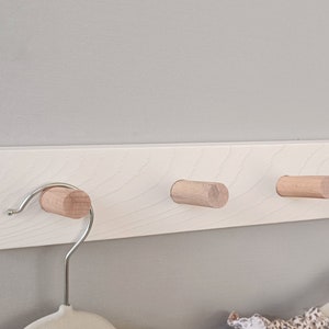 以下が含まれることがあります： 天然木製のペグと金属製のフックが付いた、明るい木製の壁掛けコートラック。ラックは灰色の壁に取り付けられています。服の付いたハンガーが2つ見えます。