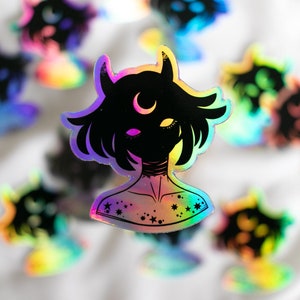 Puede incluir: Una pegatina holográfica con una ilustración en blanco y negro de una persona con cuernos y una luna creciente en la frente. La persona tiene el pelo corto y lleva un collar con estrellas. La pegatina tiene un acabado iridiscente arcoíris.