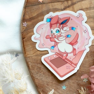 May include: A pink and white Pokemon character, Sylveon, is shown in a pink and white swirl of sparkles. Sylveon is standing on a pink Nintendo 3DS handheld gaming console.