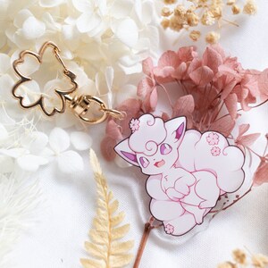 Peut inclure: Un porte-clés représentant un personnage d'anime rose et blanc avec un charme en forme de fleur dorée. Le personnage a les cheveux roses et un corps blanc et moelleux. Le porte-clés est entouré de fleurs séchées.