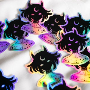 Puede incluir: Un conjunto de pegatinas hologr&aacute;ficas iridiscentes con una silueta negra de una persona con cuernos y una luna creciente. Las pegatinas tienen un acabado iridiscente arco&iacute;ris y est&aacute;n decoradas con estrellas.