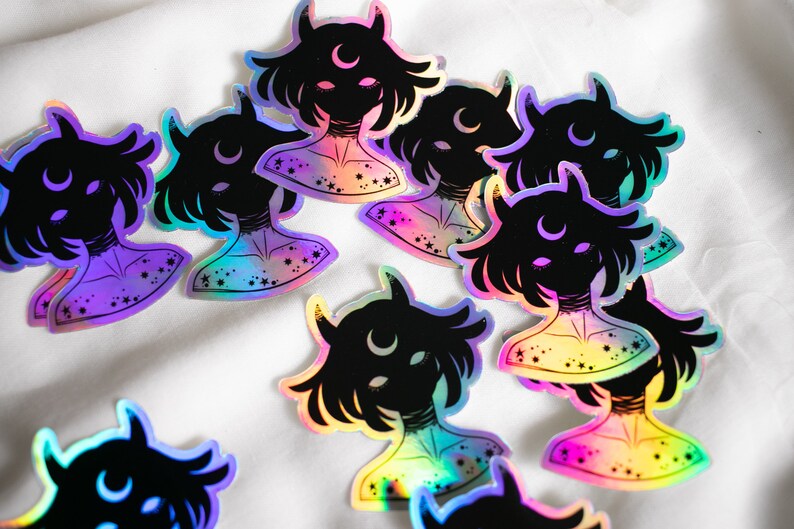 Puede incluir: Un conjunto de pegatinas hologr&aacute;ficas iridiscentes con una silueta negra de una persona con cuernos y una luna creciente. Las pegatinas tienen un acabado iridiscente arco&iacute;ris y est&aacute;n decoradas con estrellas.