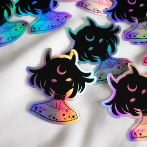 Puede incluir: Un conjunto de pegatinas hologr&aacute;ficas iridiscentes con una silueta negra de una persona con cuernos y una luna creciente en la frente. Las pegatinas tienen un acabado iridiscente arco&iacute;ris y est&aacute;n decoradas con estrellas.
