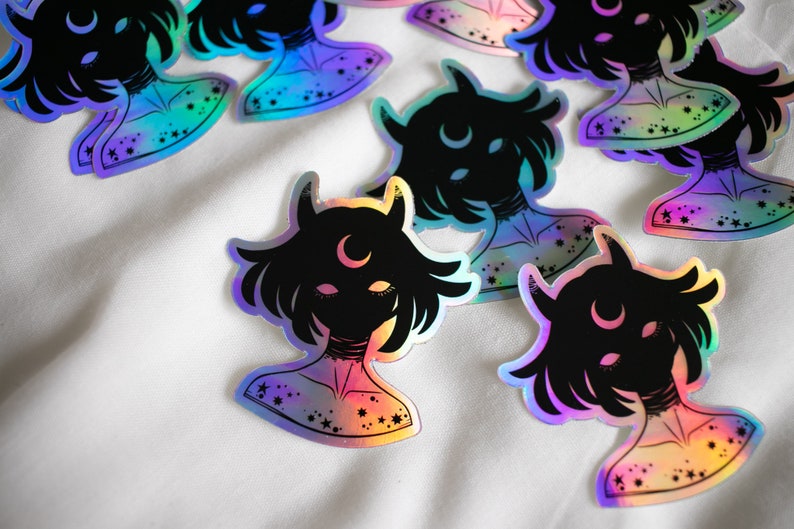 Puede incluir: Un conjunto de pegatinas hologr&aacute;ficas iridiscentes con una silueta negra de una persona con cuernos y una luna creciente en la frente. Las pegatinas tienen un acabado iridiscente arco&iacute;ris y est&aacute;n decoradas con estrellas.
