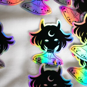Puede incluir: Un conjunto de pegatinas hologr&aacute;ficas iridiscentes con una silueta negra de una persona con cuernos y una luna creciente en la frente. La persona tiene el pelo corto y lleva un collar. Las pegatinas tienen un acabado iridiscente arco&iacute;ris y est&aacute;n decoradas con estrellas.