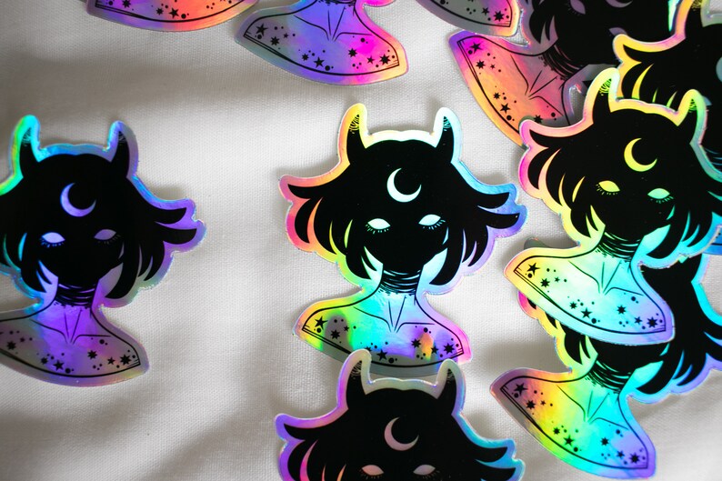 Puede incluir: Un conjunto de pegatinas hologr&aacute;ficas iridiscentes con una silueta negra de una persona con cuernos y una luna creciente en la frente. La persona tiene el pelo corto y lleva un collar. Las pegatinas tienen un acabado iridiscente arco&iacute;ris y est&aacute;n decoradas con estrellas.