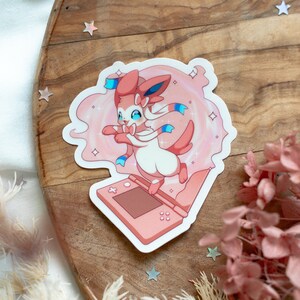 May include: Pink and white Pokemon sticker featuring a pink Sylveon with blue ribbons, holding a pink Nintendo 3DS.