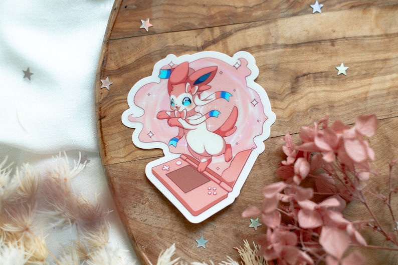 May include: Pink and white Pokemon sticker featuring a pink Sylveon with blue ribbons, holding a pink Nintendo 3DS.