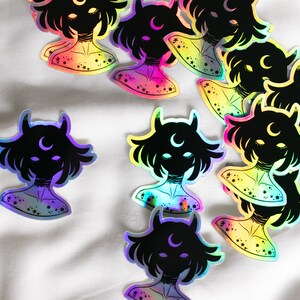 Puede incluir: Una colecci&oacute;n de pegatinas hologr&aacute;ficas iridiscentes con una silueta negra de una persona con cuernos y una luna creciente. Las pegatinas tienen un fondo degradado arco&iacute;ris con estrellas.