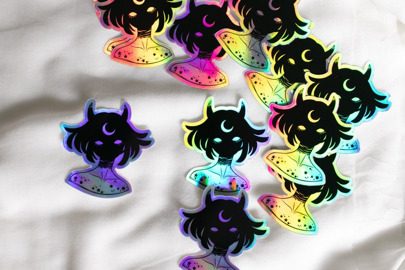 Puede incluir: Una colecci&oacute;n de pegatinas hologr&aacute;ficas iridiscentes con una silueta negra de una persona con cuernos y una luna creciente. Las pegatinas tienen un fondo degradado arco&iacute;ris con estrellas.