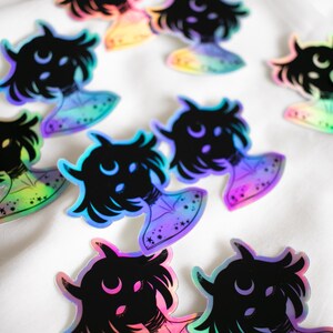 Puede incluir: Una colecci&oacute;n de pegatinas hologr&aacute;ficas iridiscentes con una silueta negra de una persona con cuernos y una luna creciente sobre su cabeza. Las pegatinas tienen un brillo arco&iacute;ris y est&aacute;n dispuestas sobre una superficie blanca.