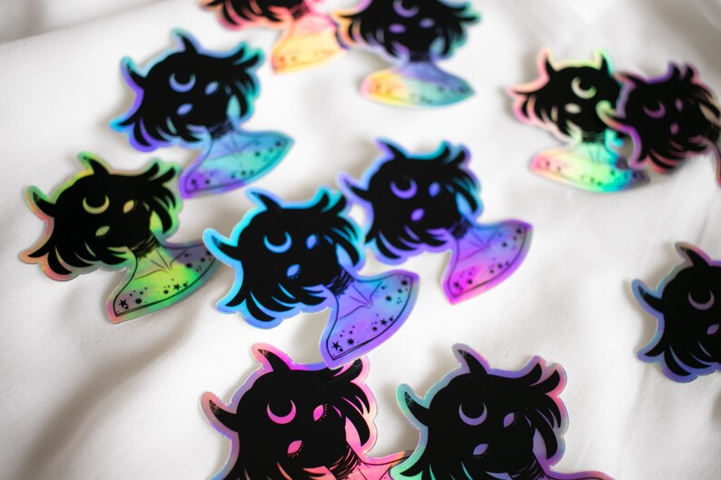 Puede incluir: Una colecci&oacute;n de pegatinas hologr&aacute;ficas iridiscentes con una silueta negra de una persona con cuernos y una luna creciente sobre su cabeza. Las pegatinas tienen un brillo arco&iacute;ris y est&aacute;n dispuestas sobre una superficie blanca.
