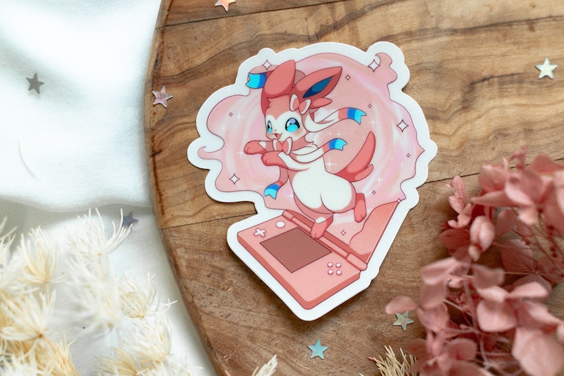 May include: A pink and white Pokemon character, Sylveon, is shown in a pink and white swirl of sparkles. Sylveon is standing on a pink Nintendo 3DS handheld gaming console.
