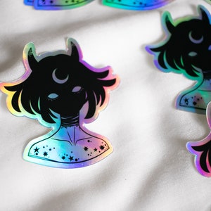 Puede incluir: Una pegatina hologr&aacute;fica con la silueta negra de una persona con cuernos y una luna creciente en la frente. La persona tiene estrellas en el pecho y el cuello. La pegatina tiene un acabado iridiscente arco&iacute;ris.