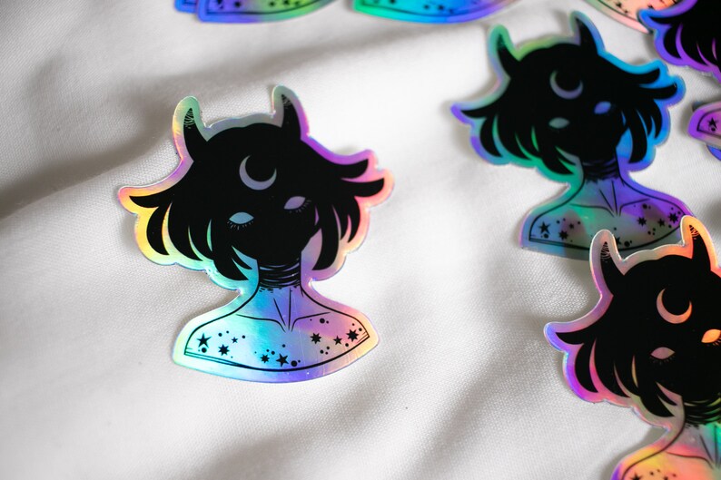 Puede incluir: Una pegatina hologr&aacute;fica con la silueta negra de una persona con cuernos y una luna creciente en la frente. La persona tiene estrellas en el pecho y el cuello. La pegatina tiene un acabado iridiscente arco&iacute;ris.
