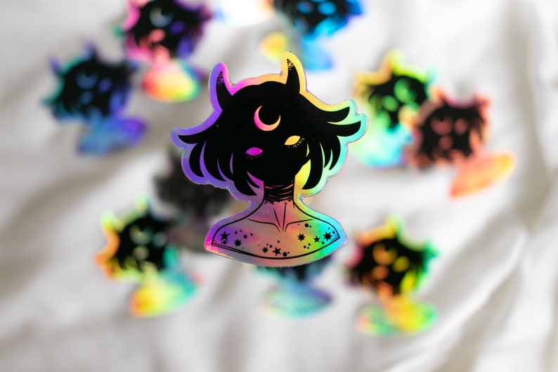 Puede incluir: Una pegatina hologr&aacute;fica con una ilustraci&oacute;n en blanco y negro de una persona con cuernos y una luna creciente en la frente. La persona tiene el pelo corto y lleva un collar con estrellas. La pegatina tiene un acabado iridiscente arco&iacute;ris.