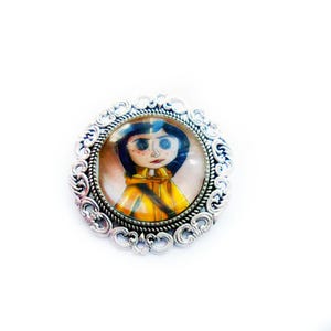 Pu&ograve; includere: Una spilla argentata con un cabochon rotondo in vetro che presenta un'illustrazione disegnata a mano di una ragazza con occhi blu e capelli neri che indossa una giacca gialla.